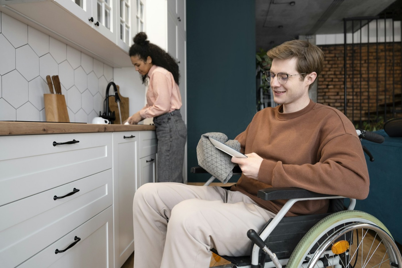 A man in a wheelchair reading at home while a woman works in the kitchen behind him